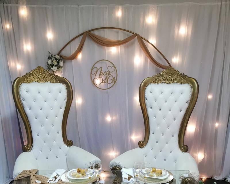 Table d'honneur dressée pour des noces d'or : deux fauteuils blancs à cadre doré devant un rideau blanc éclairé et une arche dorée